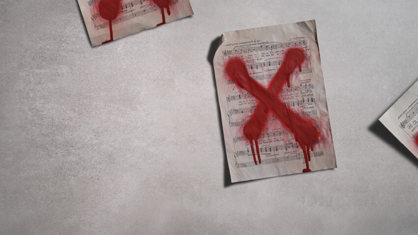 A sheet of music lies on a grey surface with a large, red, spray-painted X across it. Other music sheets marked with red paint are partially visible in the corners.